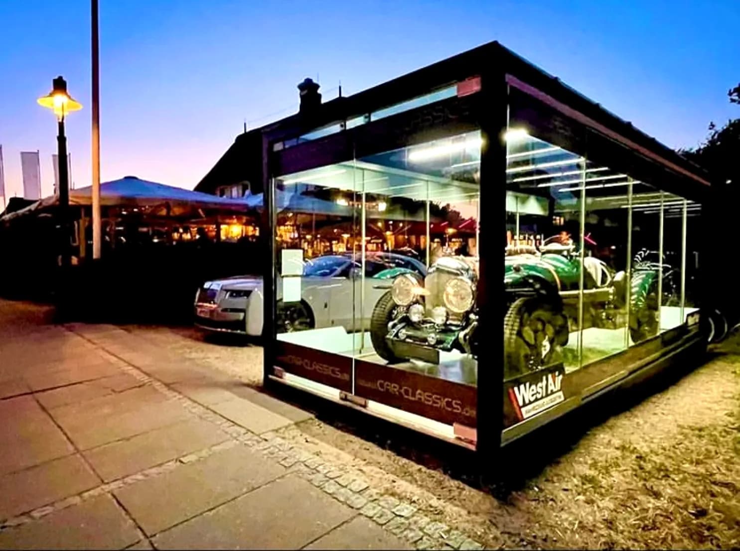 REINHART Gallery Car Classics bei Nacht: beleuchtete Glasvitrine mit Sportwagen, Licht spiegelt sich auf dem nassen Pflaster. Projektfoto.